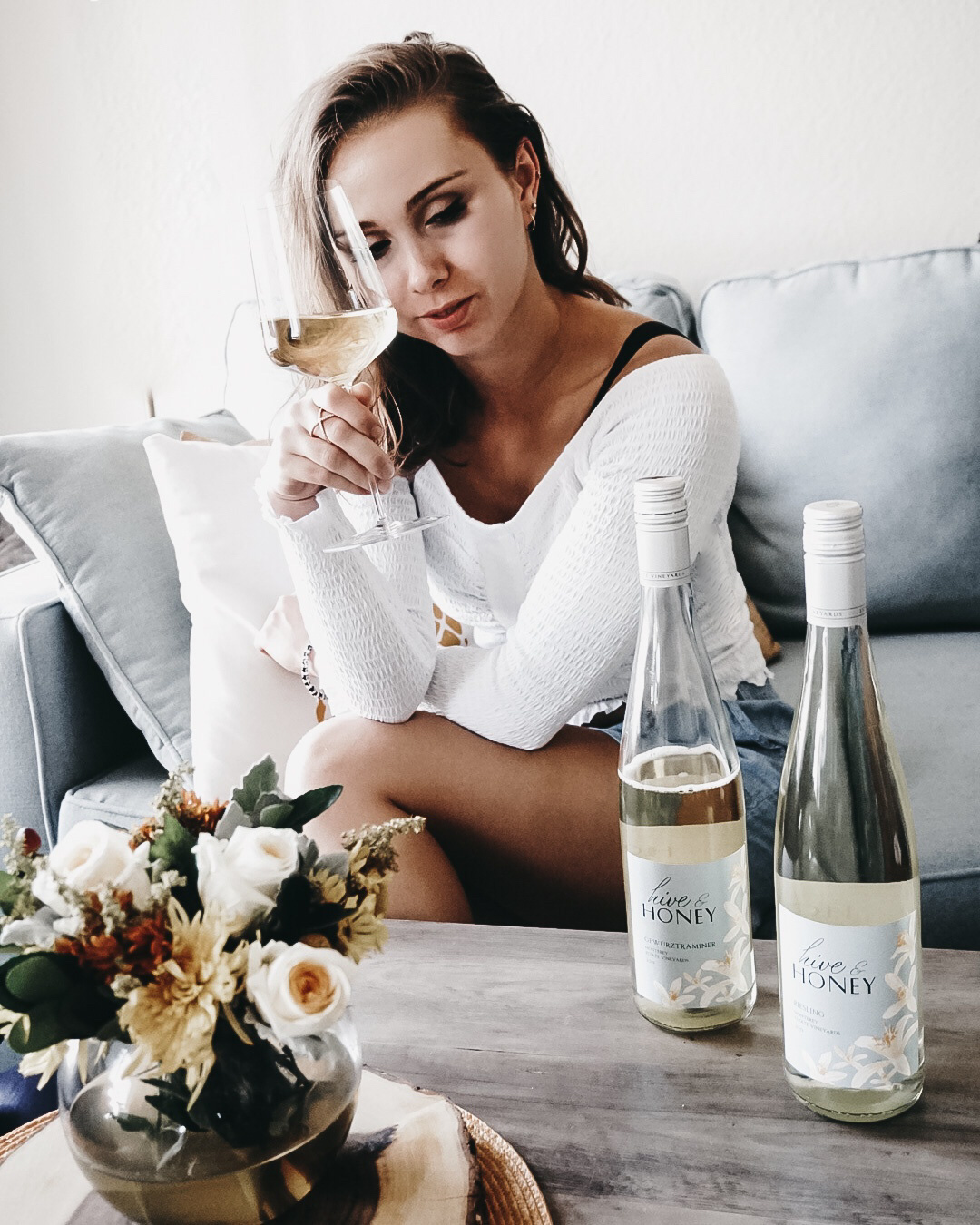 A lady sittng on a couch with two bottles of Gewurztraminer wine on a table and hold a wine glass filled with wine