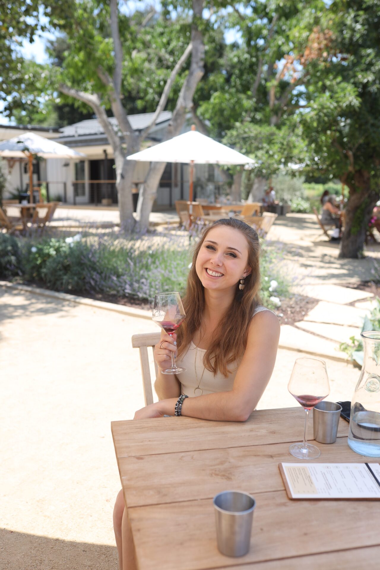 A lady sitting on a chair with a table smiling and holding a wine glass filled with wine